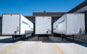 Three white cargo trailers parked at an industrial shipping dock under clear blue skies.